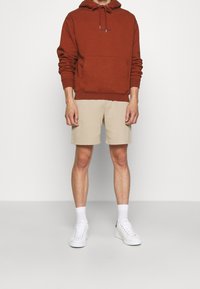 Röd bomulls-hoodie med justerbar dragsko, beige shorts och vita sneakers. Outfiten har en framficka på hoodien.