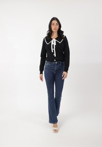 Suéter negro con un gran cuello blanco y detalle de corbata, combinado con jeans de mezclilla azul de tiro alto y acampanados, y tacones blancos destalonados.