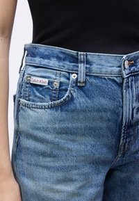 Άτομο που φοράει μπλε ψηλόμεσα τζιν τζην με ορατή ετικέτα Calvin Klein στην μικρή μπροστινή τσέπη, συνδυασμένα με μαύρο τοπ.