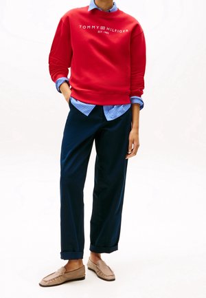 Sudadera roja con el logo "TOMMY HILFIGER EST. 1985", camisa de rayas azules y blancas debajo, pantalones azul marino y mocasines beige.