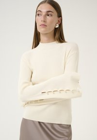 Crèmekleurige geribbelde trui met een hoge hals en lange mouwen, voorzien van parelachtige accenten langs de mouwopeningen. Gepaard met een satijnen grijze rok.