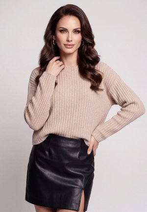 Vrouw met lang golvend haar draagt een beige geribde trui en een zwarte leren rok met een split, poseert met één hand op de heup en de andere bij haar gezicht.