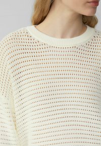 Sweter w kolorze kremowym o luźnym splocie z okrągłym ściągaczem, charakteryzujący się wzorem naprzemiennych dużych i małych otworów dla lepszej wentylacji.