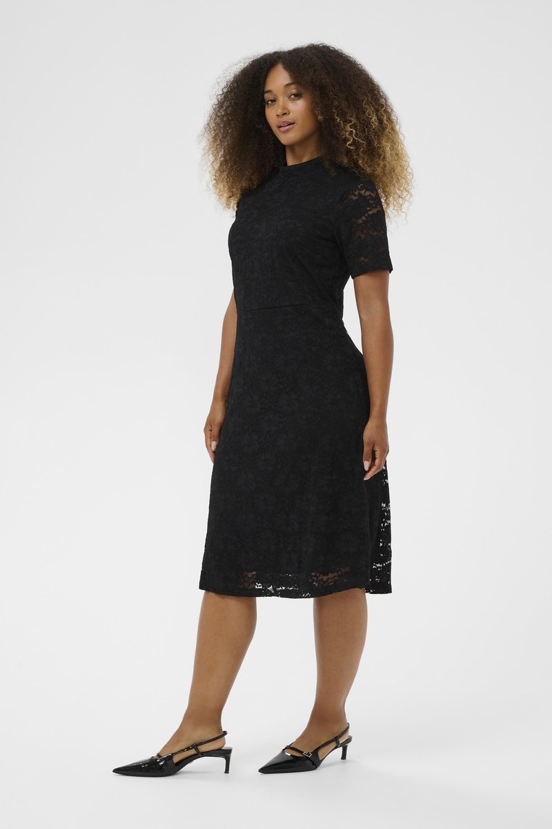 Frau mit lockigem Haar, die ein schwarzes Spitzenkleid in Knielänge und schwarze Slingback-Schuhe mit niedrigem Absatz trägt, steht vor einem einfarbigen Hintergrund.