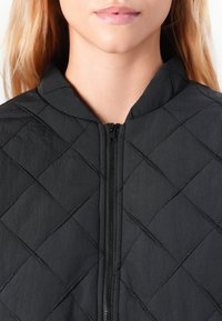 Veste matelassée noire avec col montant, dotée d'une fermeture éclair à l'avant et d'une texture en diamant. Conçue en matériau synthétique léger.