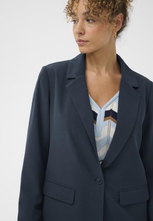 Frau mit lockigem Haar, die goldene Creolen trägt, einen marineblauen Blazer und ein V-Ausschnitt-Oberteil mit blau-weißen und braunen Mustern anhat, schaut nach rechts.