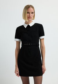 Donna con capelli biondi a caschetto, che indossa un vestito nero corto con colletto e polsini bianchi, una cintura nera e collant neri trasparenti.