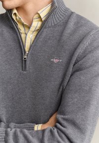 Mann trägt einen grauen Half-Zip-Pullover über einem gelb gestreiften Hemd mit Knöpfen, die Arme verschränkt, zeigt das bestickte Logo auf der Pulloverbrust.