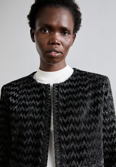 Black textured jacket with a chevron pattern, featuring a round neckline and front closure. Made with a shimmering fabric, interior is light.