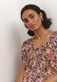 Bluse med blomstermotiv i pink, lilla og grønne farver. Blød stof, løs pasform, med nøglehulkrave og samlede korte ærmer.
