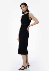 Czarna sukienka bez rękawów o dopasowanym kroju i pasku w talii. Gładka tekstura, do kolan, zestawiona z czarnymi sandałkami na obcasie.