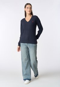 Maglione navy a trecce con scollo a V e polsini a costine, abbinato a jeans ampi in lavaggio chiaro e sneakers bianche. Dettaglio del logo rosso sul maglione.