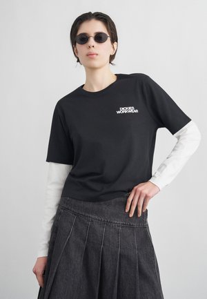 Osoba ubrana w czarny T-shirt "Dickies Workwear" na białych długich rękawach, ciemną plisowaną spódnicę i małe owalne okulary przeciwsłoneczne, pozująca z ręką na biodrze.