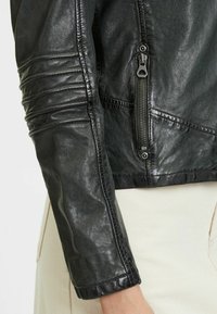 Zwarte leren jas met een gestructureerde afwerking, aangesloten model, ritssluiting en gestikte details op mouwen en lijf.