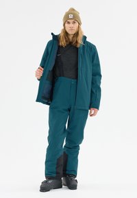 Teal vattentät jacka och byxor, med fickor, dragkedjor och svarta accenter. Bärs över en grå tröja med en beige mössa.