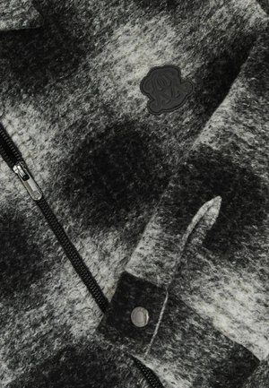 Šedá a bílá fleecová bunda se zipovým uzávěrem, texturovaným vzorem a černou nášivkou na hrudi. Ozdobena patentkami.