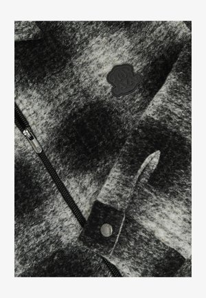 Szürke és fehér polár dzseki, cipzáros záródással, texturált mintázattal és fekete logóval a mellkason. Nyomógombokkal díszítve.