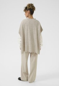 Poncho en tricot beige avec bordure festonnée, porté par-dessus un haut à manches en dentelle blanche et assorti à un pantalon beige clair et des chaussures noires.