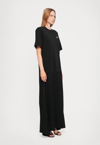 Black oversized t-shirt dress with short sleeves, smooth texture, and a slight high-low hem. Features a small logo detail on the chest.