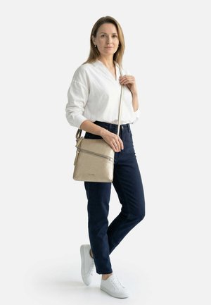 Žena v bílé blůze, tmavých kalhotách a bílých teniskách, držící béžovou kabelku přes rameno s předním zipovým kapsou, na jednobarevném pozadí.