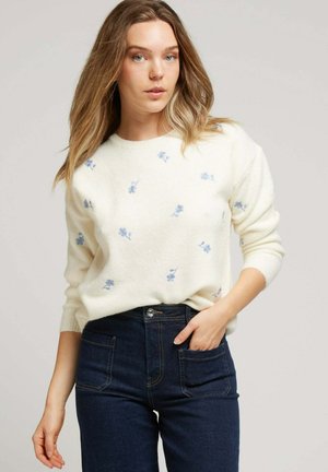 Pull en tricot blanc avec de la broderie florale bleue, col rond et manches longues, associé à un jean en denim foncé taille haute.