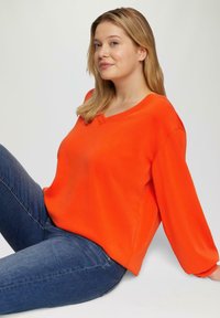 Helles orangefarbenes V-Ausschnitt-Pullover mit langen, lockeren Ärmeln und glatter Textur, kombiniert mit blauen Denim-Jeans vor einem neutralen Hintergrund.