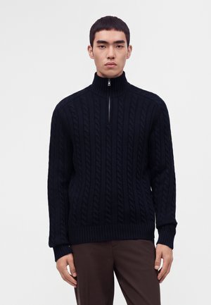 Jeune homme portant un pull en maille torsadée noire avec une courte fermeture éclair et un pantalon marron, debout devant un fond blanc uni.