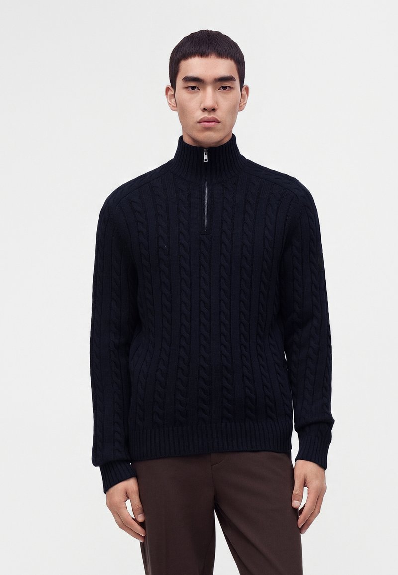 Jeune homme portant un pull en maille torsadée noire avec une courte fermeture éclair et un pantalon marron, debout devant un fond blanc uni.