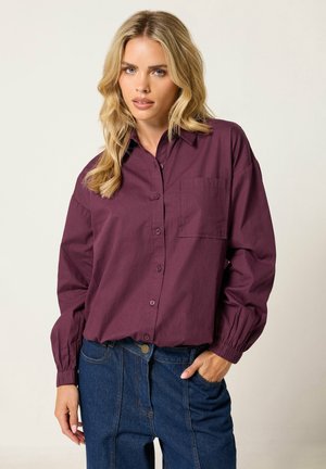 Femme blonde portant une chemise violette à manches longues avec une poche poitrine et un jean taille haute bleu foncé, debout avec une main dans la poche.