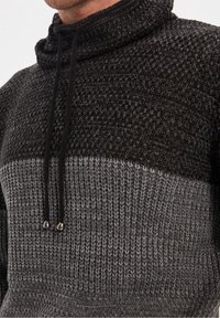 Gebreide pullover met een hoge kraag, met een textuurpatroon in zwart en grijstinten, en verstelbare trekkoordjes met metalen uiteinden.