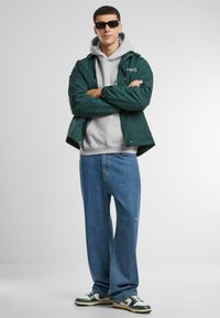 Green jacket, gray hoodie, loose blue jeans, and green and white sneakers; model wearing black sunglasses. Smooth textures and casual style.