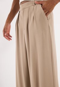 Béžové kalhoty s širokými nohavicemi vyrobené z hladkého materiálu; mají přehyby, boční kapsy a upravený pas pro elegantní vzhled.