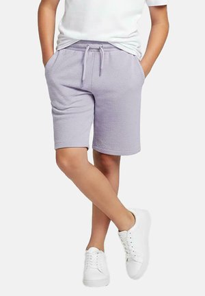 Persoon met lichtpaarse short met trekkoord en handen in de zakken, wit t-shirt en witte sneakers, staand met gekruiste benen.