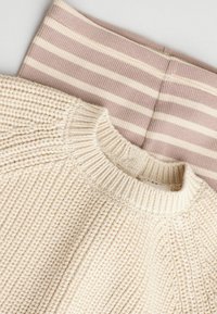 Pull en tricot beige clair avec un col côtelé, superposé sur des leggings rayés mauve clair, avec des rayures crème alternées.