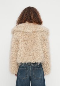 Άτομο με καστανά σγουρά μαλλιά, που φοράει μια μπεζ τζάκετ από χνουδωτή γούνα και σκούρα μπλε ψηλόμεσα τζιν, εμφανίζεται από πίσω σε απλό φόντο.