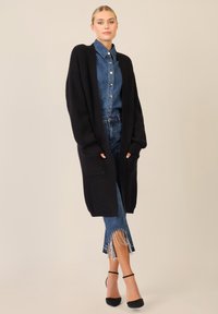 Cardigan lungo nero a maglia con apertura frontale e tasche, abbinato a una camicia di jeans blu e jeans con orlo frangiato, che completano sandali neri con tacco.