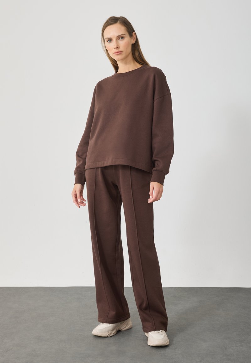 Brązowy dres składający się z bluzy i spodni, wykonany z miękkiego materiału. Luźny krój, okrągły dekolt i długie rękawy, z prążkowanymi mankietami i szwami.