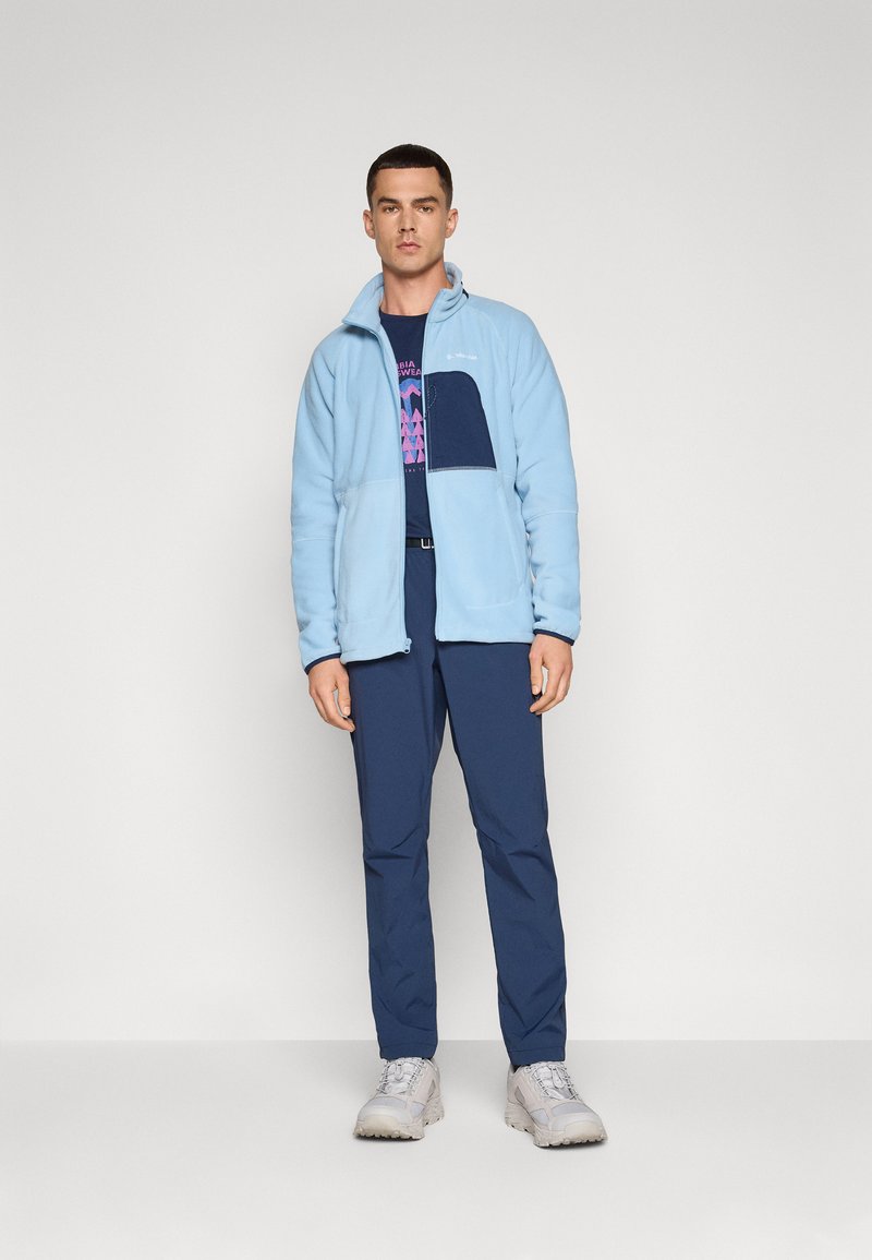 Jasnoniebieska polarowa kurtka z granatową kieszenią, noszona na ciemnej koszuli. W zestawie z granatowymi spodniami i jasnoszarymi sneakersami.
