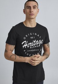 Crna pamukasta majica s kratkim rukavima, s velikim bijelim grafičkim tekstom koji glasi "Heritage Blend" i ostalim detaljima brendiranja.