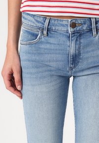 Jean en denim bleu clair au design ajusté, avec des poches arrière, un délavage léger et un bouton en bronze à la taille.