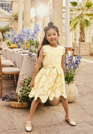 Jeune fille en robe jaune en dentelle et chaussures dorées se tenant dans une pièce éclairée par le soleil avec une longue table et des arrangements floraux bleus.