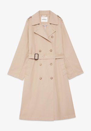 Kvinde iført en beige trenchcoat over en grøn og hvid blomsterdragt med vidde bukser og beige flade sko, stående mod en ensfarvet baggrund.