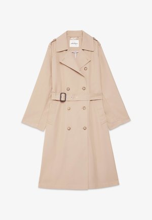 Vrouw draagt een beige trenchcoat over een groen-wit gebloemd pak met wijde pijpen en beige platte schoenen, staand tegen een effen achtergrond.