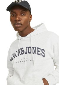 Sudadera gris clara con un bolsillo canguro frontal y capucha con cordón. Presenta texto y acentos en azul marino oscuro. Se lleva con una gorra azul marino.