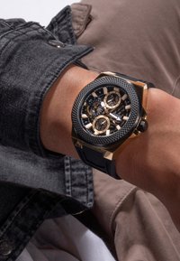 Montre chronographe noire et dorée avec lunette texturée, trois sous-cadrans et un bracelet en caoutchouc noir sur un poignet, assortie à une veste sombre.