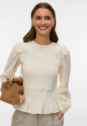Krēmkrāsas blūze ar strukturētu šuvi, garām pūkainām piedurknēm un volāniem pie apakšas. Kombinēta ar brūnu zamša soma.