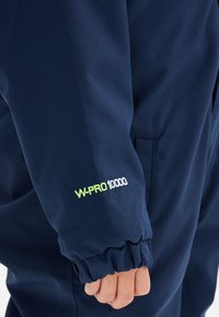 Marineblauwe waterdichte jas met elastische manchetten; bevat het "W-PRO 10000" logo in wit en groen op de mouw.