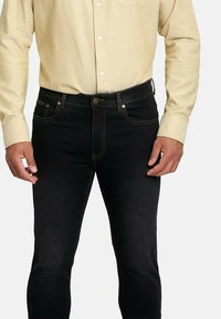 Jeans de mezclilla negra con corte ajustado, con costuras amarillas, bolsillos frontales y cierre de un solo botón. Llevados con una camisa de color claro.