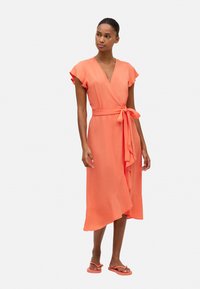 Vestido envolvente coral con mangas cortas de volantes, un cinturón atado a la cintura y un dobladillo asimétrico y fluido. Llevado con chanclas a juego.