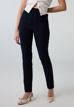 Vrouw draagt nauwsluitende zwarte broek, crème mouwloos topje met knopen en crème slingback-hakken met strikken, staand tegen een effen achtergrond.